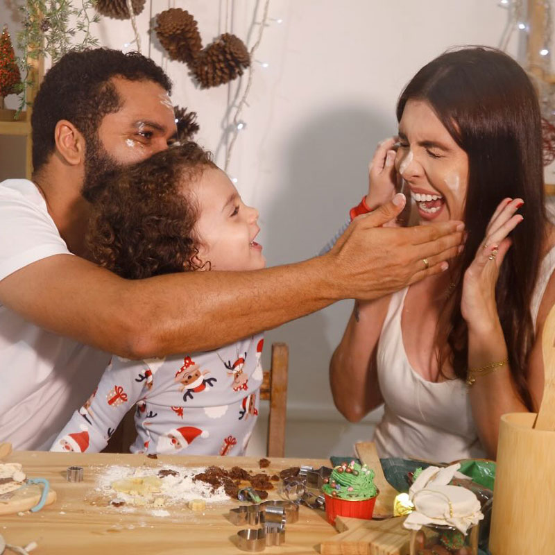 Parents building a gingerbread house with their young child. The father is smearing icing on the mothers face while they all laugh