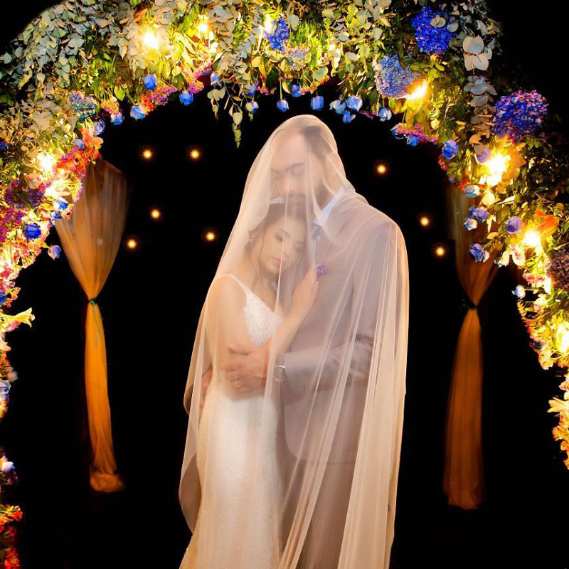 Newlywed couple poses under floral arch. The bride's veil covers them both
